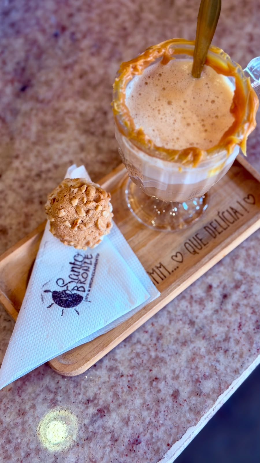 Cappuccino com calda de caramelo servido em taça, ao lado de um docinho, em bandeja de madeira com a marca Santo Bronze.