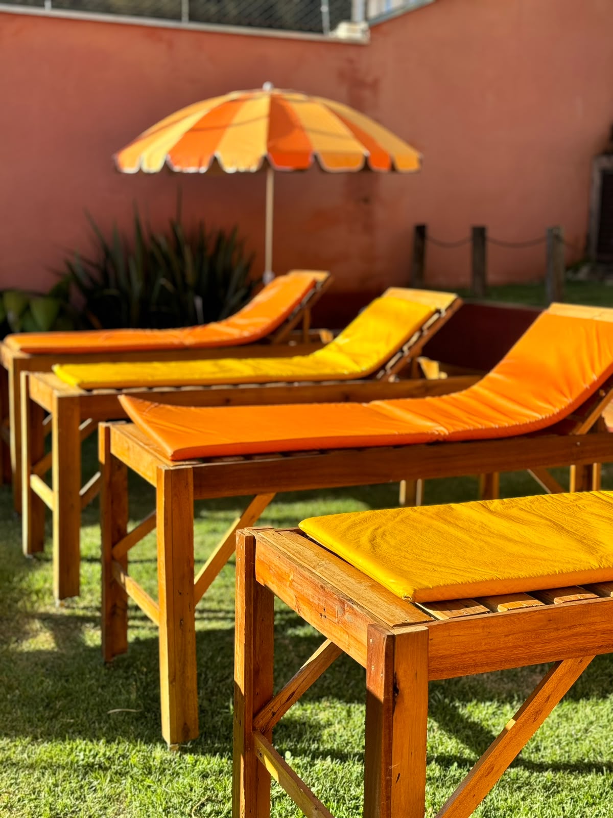 Espreguiçadeiras de madeira com almofadas laranja e amarela, guarda-sol e plantas ao fundo.