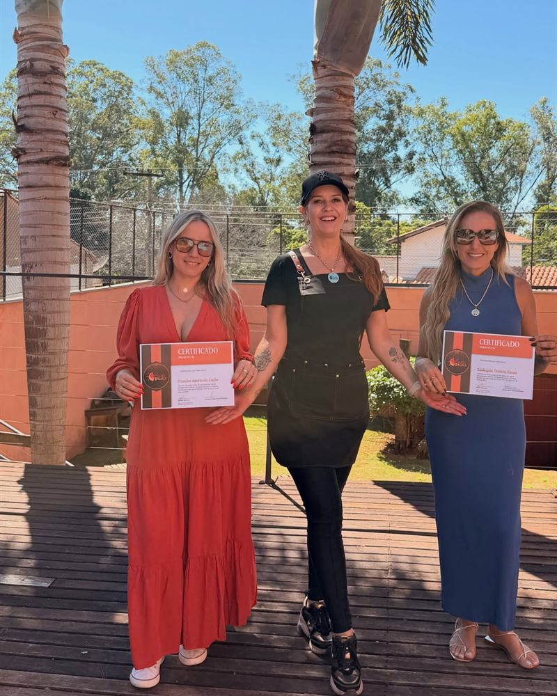 Diana Alice ao ar livre entre duas alunas que seguram seus certificados do curso de bronzeamento.
